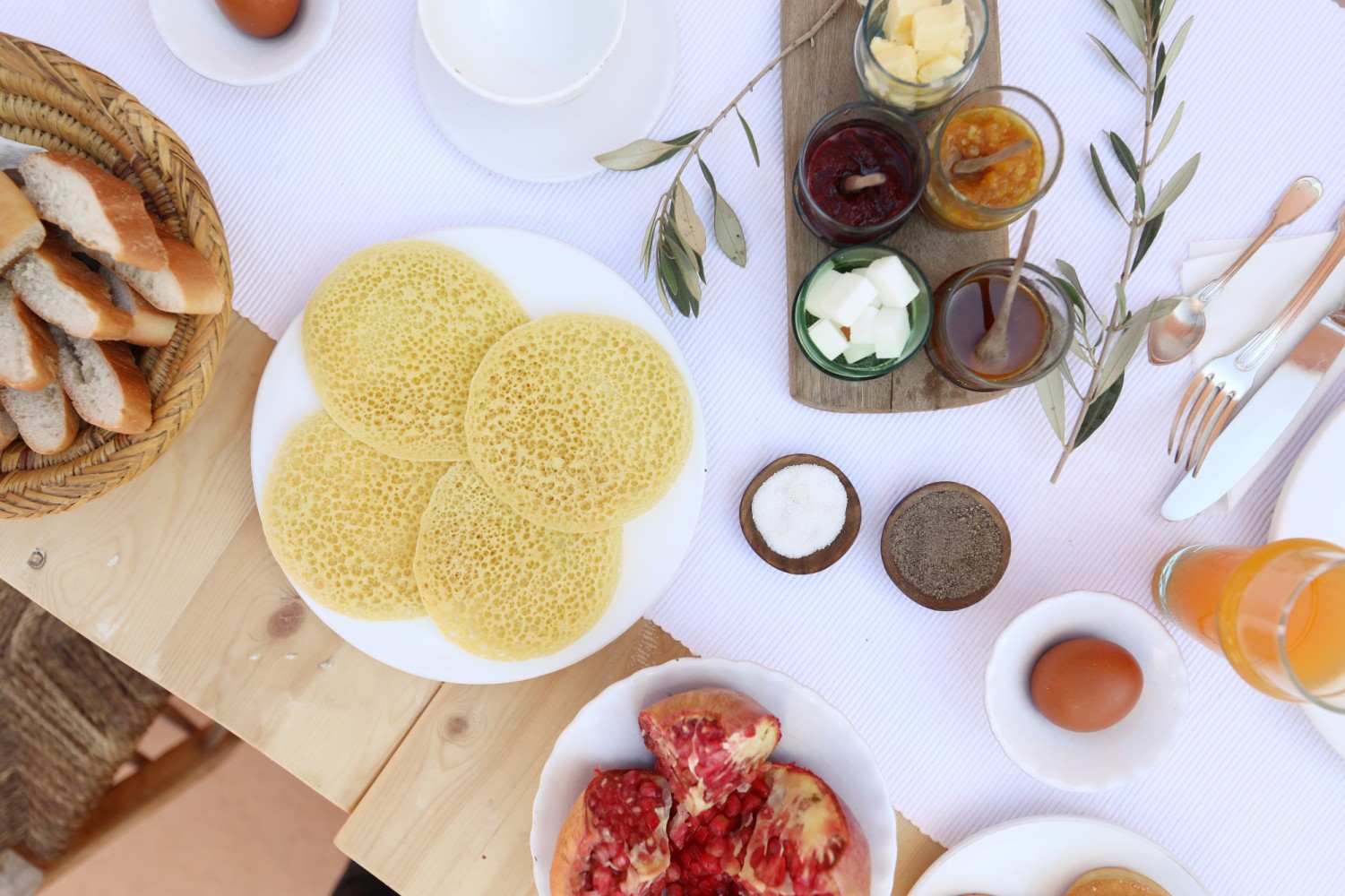 La table du petit-déjeuner vue de haut — beghrir, corbeille de pain, confitures, œufs, jus frais et grenade