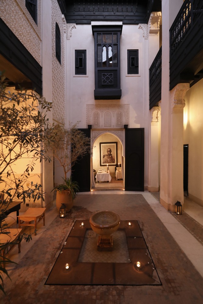 Le patio du riad au crépuscule, la porte du salon éclairée derrière les oliviers